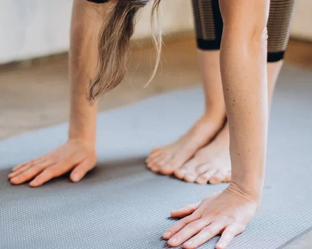A person in a yoga pose with a close up of hands and bare feet on a grey-blue yoga mat.