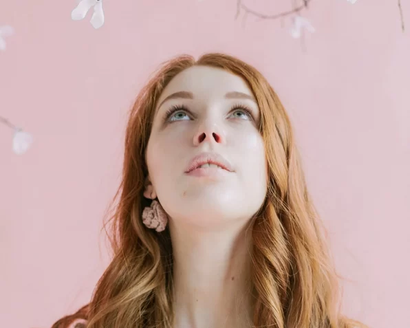 A headshot of a youngish woman with long ginger hair, looking upwards towards floral decorations.